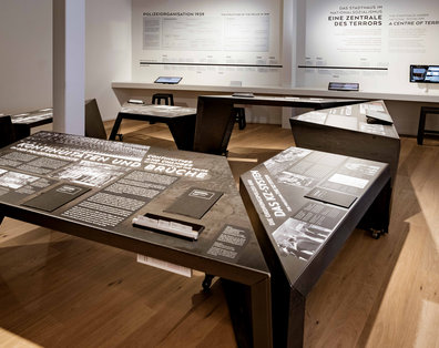 Blick in die Ausstellung am Geschichtsort Stadthaus auf einen großen dunklen Ausstellungstisch mit Fotos und Texten.